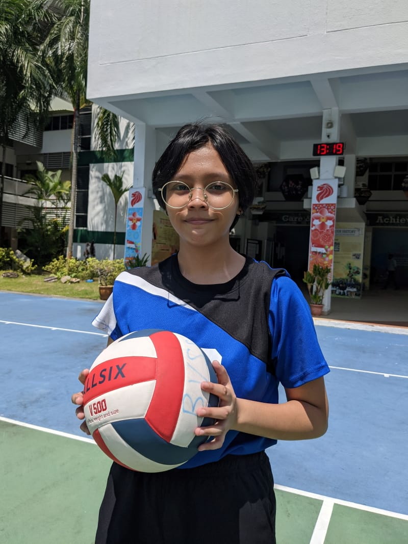 Young girl with glasses in a blue sports jersey holds a red and white volleyball on an outdoor court.